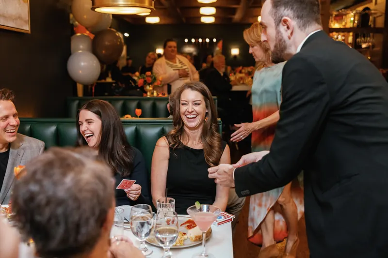 Wedding guests laughing at a close-up card trick performed tableside during the reception